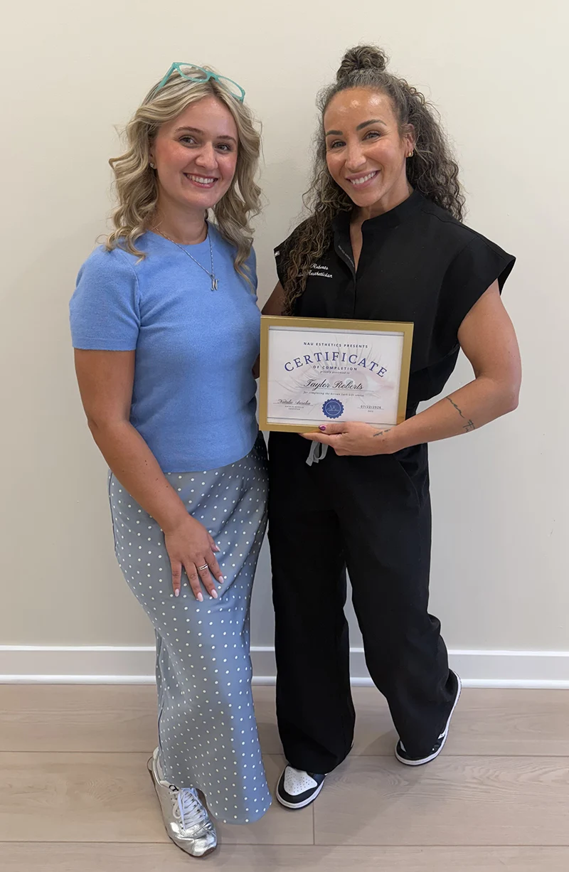 Aesthetician holding a framed training certificate of completion alongside her instructor.